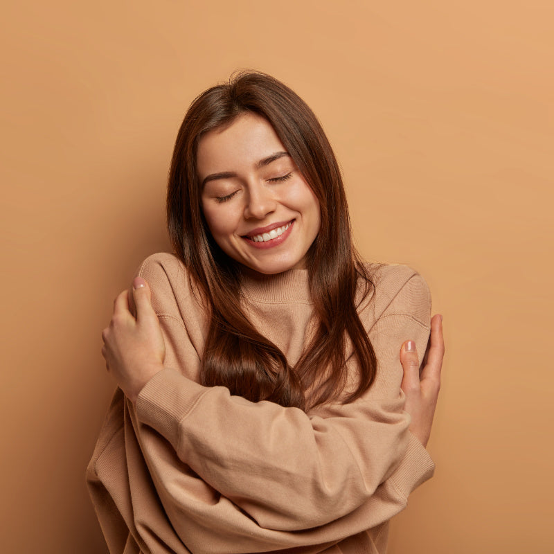 A split-screen image with a warm beige background. On the left, a smiling woman with long brown hair, wearing a beige sweater, hugs herself with closed eyes, conveying a sense of comfort and well-being. On the right, black text on a white background reads: 'REASON 1 - Gentle & Effective. Coconut-derived surfactants (SCI and SCS) create a rich lather that cleans thoroughly without stripping hair of its natural oils.'